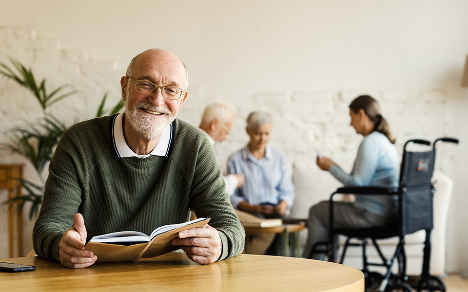 Senior man smiling while reading a book sitting at table in an assisted living facility